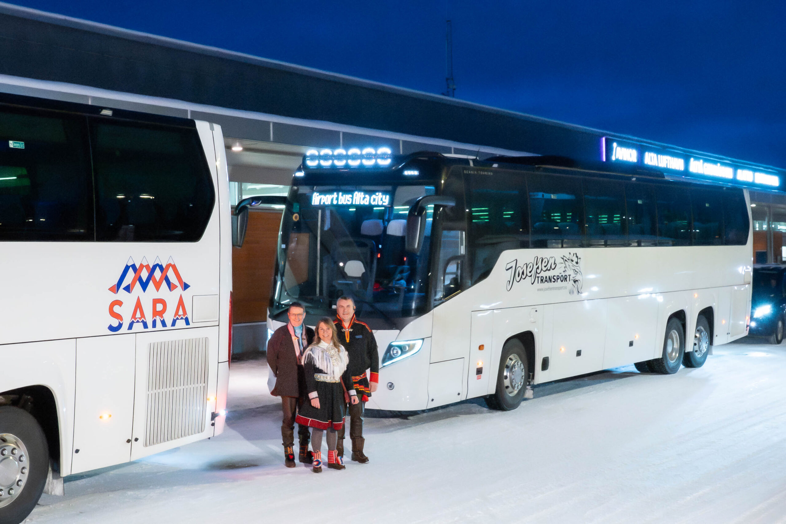 Tre samer i kofte står foran M.M. Sara transport og Josefsens transports busser ved Alta flyplass